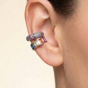 Puede incluir: Dos puños de oreja con piedras preciosas de colores. El puño superior está cubierto de pequeñas piedras multicolores. El puño inferior presenta una fila de piedras rectangulares de colores arcoíris. La joya se lleva en la oreja.