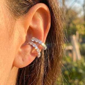 Puede incluir: Primer plano de unos pendientes de oreja con una fila de piedras preciosas claras y brillantes. Los pendientes son de color plateado y se curvan alrededor de la oreja. La imagen destaca el diseño de la joya y la forma de la oreja.