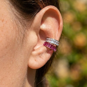 Puede incluir: Pendiente de oreja con dos bandas de piedras preciosas. La banda superior tiene piedras claras, de corte rectangular, y la banda inferior piedras rosas vibrantes, de corte rectangular. La joya se lleva en la oreja, mostrando un diseño moderno y elegante.