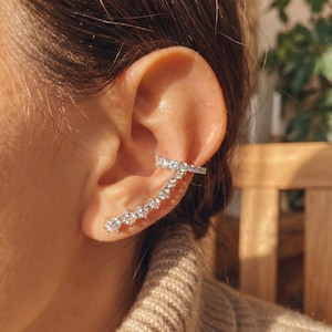 May include: A silver ear cuff with a series of clear, sparkling stones. The cuff curves along the ear, with a bar that wraps around the upper ear and a line of stones that cascade down the earlobe. The jewelry is set against a blurred background.