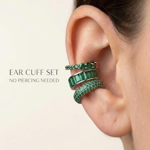 May include: A set of three silver-toned ear cuffs featuring green stones. The top cuff has round stones, the middle has baguette-cut stones, and the bottom has pavé-set stones. Text reads: EAR CUFF SET NO PIERCING NEEDED. These cuffs sit on the ear cartilage.