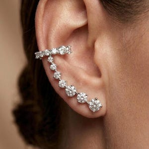 Può includere: Un ear cuff color argento caratterizzato da una fila di pietre rotonde, trasparenti e sfaccettate che avvolgono la cartilagine superiore e scendono lungo il bordo esterno dell'orecchio, con ogni pietra fissata in una montatura a griffe.