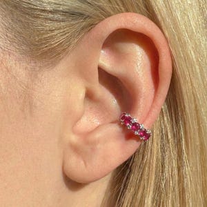 May include: A silver ear cuff adorned with vibrant red gemstones and small, sparkling white stones. The cuff curves along the ear's cartilage, creating a delicate and eye-catching accessory. The gemstones are evenly spaced.