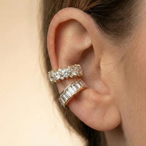 May include: Two gold ear cuffs worn on the cartilage. The top cuff features a row of round brilliant-cut stones, while the bottom cuff displays a channel-set row of rectangular baguette-cut stones, both set in a polished gold-tone metal band.