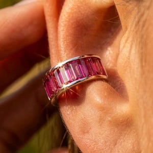 Può includere: Un orecchino a cerchio d'argento con una fila di pietre preziose rosa rettangolari e vibranti. L'orecchino è indossato sull'orecchio, mettendo in risalto il colore brillante e accattivante delle pietre. Il gioiello è progettato per essere indossato senza piercing.