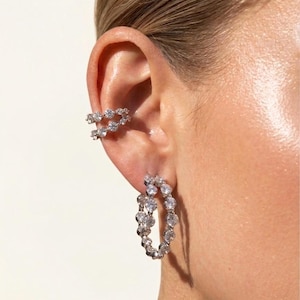 Könnte beinhalten: Nahaufnahme eines Ohrs mit funkelndem Schmuck. Das Ohr zeigt einen doppelreihigen Ohrklemmen-Aufsatz und ein Paar Creolen, beide mit klaren, runden Edelsteinen besetzt. Der Schmuck glänzt auf der Haut.