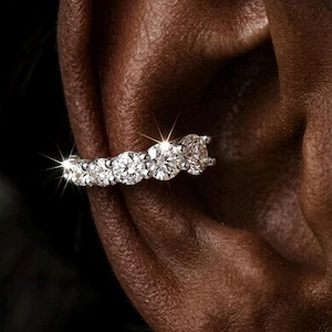 May include: A silver-toned ear cuff featuring a row of five round-cut, clear, sparkling stones set in a metallic band. The jewelry piece is positioned along the outer edge of the ear cartilage, showcasing a bright, reflective, and faceted finish.