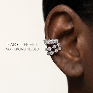 Op de afbeelding: Een zilverkleurige ear cuff set bestaande uit drie gestapelde ringen bezet met heldere, rondgeslepen stenen. De tekst "EAR CUFF SET NO PIERCING NEEDED" staat links van het sieraad gedrukt. Het ontwerp klemt om de rand van het oor.