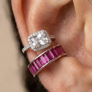 May include: Two silver-toned ear cuffs worn on the ear cartilage. The top cuff features a rectangular clear stone surrounded by a halo of small white stones. The bottom cuff is a band set with a row of rectangular vibrant pink stones.