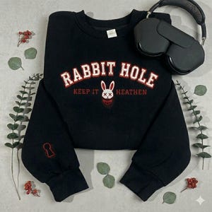 May include: Black crewneck sweatshirt with the text "RABBIT HOLE" in white and red lettering, a bunny graphic, and the words "KEEP IT HEATHEN". A keyhole graphic is on the sleeve. Black headphones rest on the sweatshirt.
