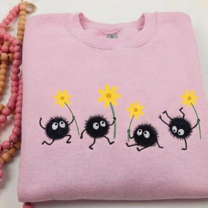 May include: A light pink sweatshirt featuring a whimsical design of four black, round characters with white eyes, holding yellow flowers. The sweatshirt has a crew neck and a soft, comfortable appearance.