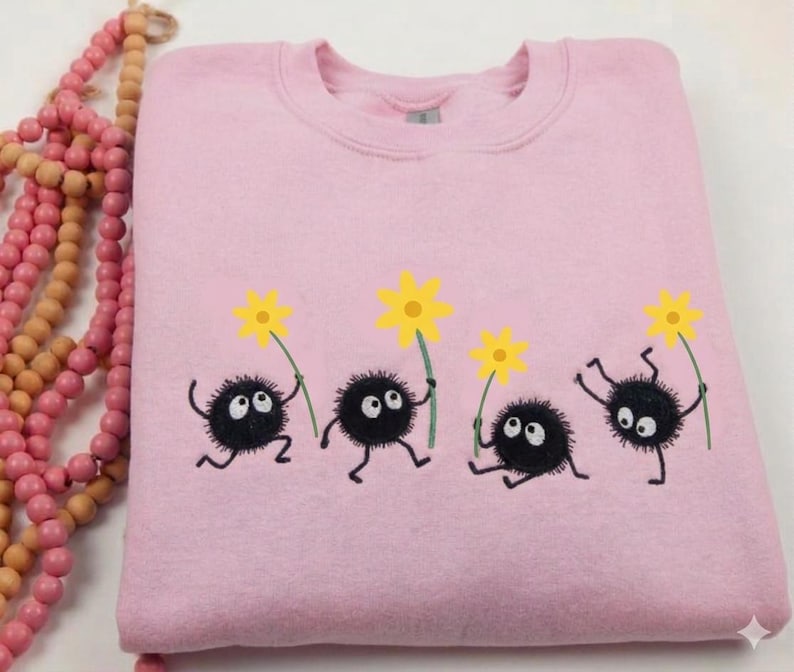 May include: A light pink sweatshirt featuring a whimsical design of four black, round characters with white eyes, holding yellow flowers. The sweatshirt has a crew neck and a soft, comfortable appearance.
