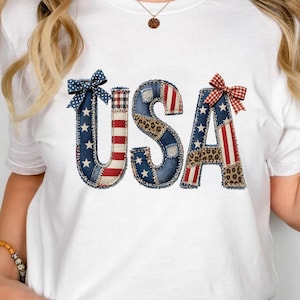Puede incluir: Una camiseta blanca con las letras USA en un diseño de patchwork. Las letras están compuestas por tela azul con estrellas blancas, rayas rojas y blancas y patrones de leopardo marrón. Un lazo azul con lunares y un lazo rojo a cuadros están unidos.