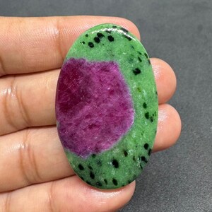 May include: An oval ruby zoisite gemstone. The stone has a central, deep magenta ruby, encircled by a vibrant green zoisite matrix. Small black spots are scattered across the green surface, providing contrast.