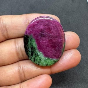 May include: Polished, oval gemstone in shades of magenta, green, and black. The stone is held in a hand, with a dark background. The gemstone's colours are vivid and well-defined.