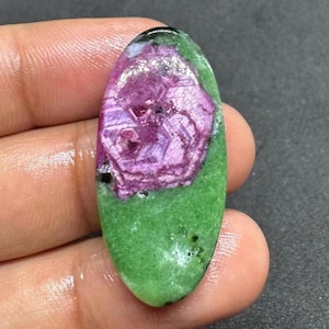 Op de afbeelding: Een ovaalvormige edelsteen met een levendige kleurencombinatie. Het midden toont een opvallende, veelzijdige, diep magenta tint, omgeven door een levendige groene basis. De steen wordt in een hand gehouden.