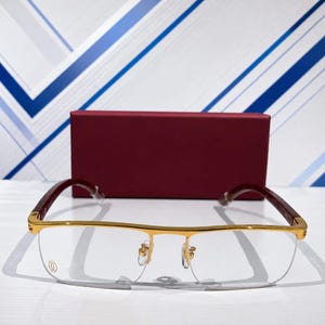 Puede incluir: Gafas semialaire doradas con montura rectangular, puente dorado fino y patillas con acabado marrón rojizo oscuro. Las lentes transparentes están montadas en una estructura de metal dorado, con un estuche rectangular color burdeos detrás.