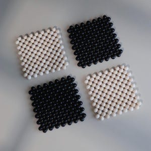May include: A set of four square coasters arranged in a grid pattern. Two coasters are composed of matte white spherical beads, and two coasters are composed of matte black spherical beads, all linked together to form a flexible, textured surface.