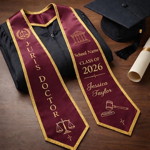 May include: Burgundy graduation sash with gold trim and text "JURIS DOCTOR" and "School Name CLASS OF 2026 Jessica Taylor". Includes a graduation cap and diploma. The sash features law-related graphics.