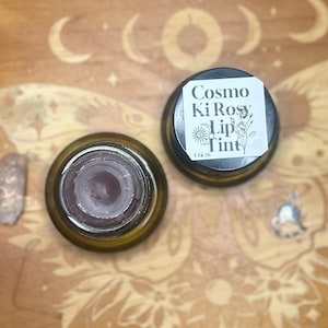 May include: An overhead shot of two small, round, brown glass jars of lip tint. One jar is open, revealing the dark red tint inside. The other jar has a black lid with a label that reads "Cosmo Ki Rosy Lip Tint."