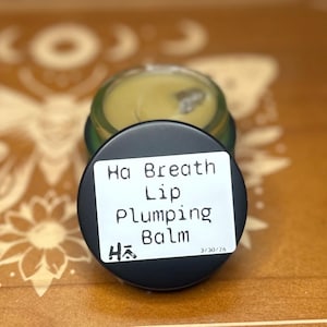 May include: A small, open jar of lip balm with a black lid. The lid has a white label with the text "Ha Breath Lip Plumping Balm." The balm is light green. The jar sits on a wooden surface.