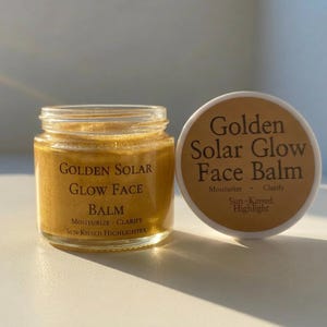 Puede incluir: Un frasco de vidrio transparente lleno de un bálsamo facial dorado, con una etiqueta que dice Golden Solar Glow Face Balm, Moisturize - Clarify, Sun-Kissed Highlighter. Al lado hay una tapa blanca con el mismo texto en marrón oscuro.