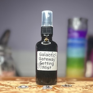 Puede incluir: Una botella de vidrio marrón oscuro con una etiqueta blanca que dice "Galactic Gateway Setting Mist". La botella tiene una tapa negra y una boquilla de pulverización transparente. El fondo está borroso, con toques de morado y verde.