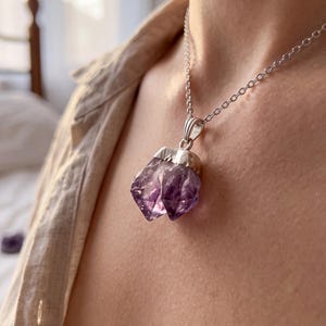 May include: A necklace featuring a raw, deep purple amethyst crystal pendant with a jagged, multi-faceted surface. The crystal is secured in a polished silver-toned metal cap and hangs from a delicate, matching silver-toned link chain.