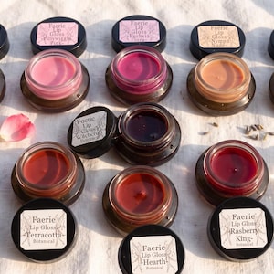 May include: A collection of ten small round glass jars containing various shades of lip gloss. Each jar has a black lid with a white label reading Faerie Lip Gloss followed by the specific shade name such as Pink Blush, Witchcraft, or Clear Glass.