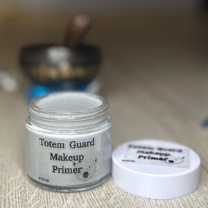 Puede incluir: Un frasco de vidrio transparente de "Totem Guard Makeup Primer" con tapa blanca. El frasco está lleno de una sustancia gris claro. La etiqueta presenta una ilustración de oso. Un fondo borroso incluye un cuenco tibetano.