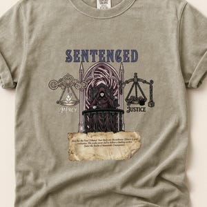May include: A light olive green t-shirt with a graphic design featuring the word "SENTENCED" in purple above an illustration of a robed figure seated in a gothic archway. The design includes scales labeled "MERCY" and "JUSTICE".