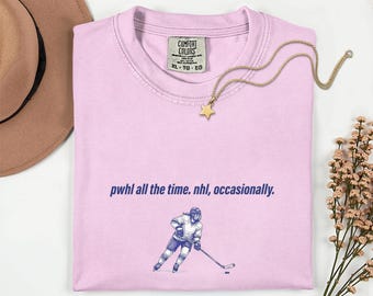 May include: A light pink short-sleeve t-shirt with a Comfort Colors label at the neck. The shirt features the dark blue text "pwhl all the time. nhl, occasionally." above a graphic illustration of a hockey player in a skating pose.