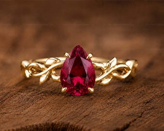 May include: A gold ring featuring a pear-shaped red gemstone set in the center. The band is crafted with a detailed vine and leaf motif that extends from the central setting, wrapping around the finger with a polished yellow gold finish.
