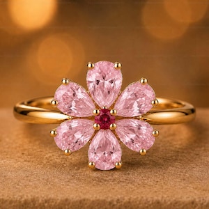 May include: A gold ring featuring a flower-shaped arrangement of five pear-cut light pink gemstones surrounding a single, small, round, deep red gemstone at the center. The band is smooth and polished, crafted from a yellow-toned metal material.
