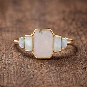 May include: A gold ring featuring a large, central, rectangular-cut moonstone flanked by two smaller rectangular moonstones and two tiny, round, iridescent opal-like stones on each side, all set in a polished gold band with a smooth finish.