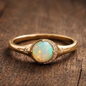 May include: A gold ring with a large, round opal center stone, surrounded by small diamonds. The band features starburst designs on each side of the opal. The ring is set against a rustic wooden background.
