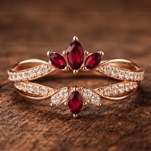 May include: A rose gold ring featuring a split band design adorned with small clear stones. The ring is accented with four marquise-cut deep red gemstones arranged in a crown-like formation at the top and a single matching red stone at the base.