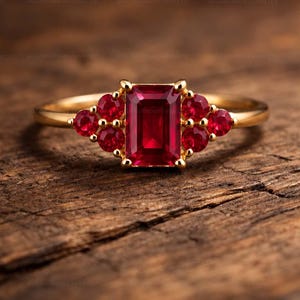 Könnte beinhalten: Ein Goldring mit einem zentralen, smaragdgeschliffenen roten Rubin, der auf jeder Seite von drei kleineren, rund geschliffenen roten Rubinen flankiert wird. Der Ring ist schmal und aus poliertem Gold gefertigt und hält die sieben Edelsteine.