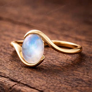 May include: A gold ring featuring an oval moonstone with a blue and white iridescent sheen. The ring's band has a flowing design that cradles the stone. The ring is displayed on a wooden surface. The moonstone is a beautiful gemstone.