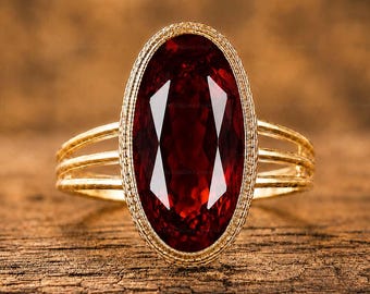May include: A gold ring featuring a large, oval-cut deep red garnet gemstone set within a textured, milgrain-detailed bezel. The band consists of three thin, parallel gold wires that connect to the central setting on each side of the stone.