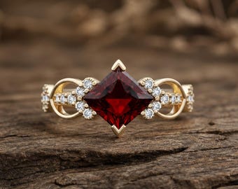 May include: A gold ring featuring a large, square-cut, deep red gemstone as the centerpiece. The ring is adorned with smaller, clear gemstones and decorative gold filigree. The ring is set on a wooden surface.