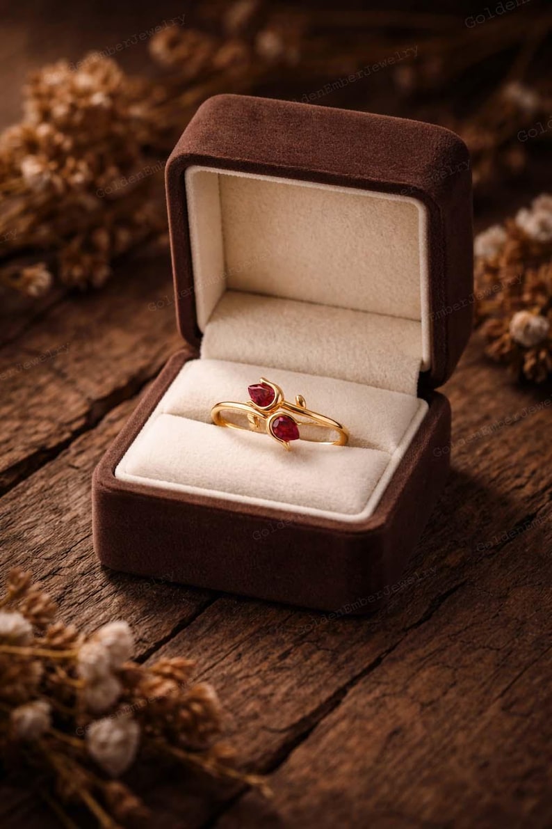 May include: A gold ring with two red gemstones, presented in a brown velvet jewelry box. The ring has a delicate, floral design. The box is open, revealing the ring on a cream-colored cushion, set against a rustic wooden background.