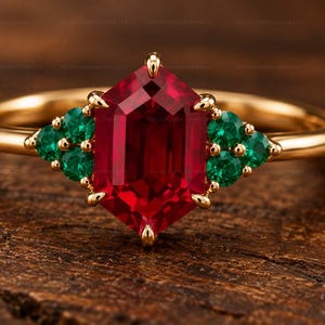 Puede incluir: Un anillo de oro con una piedra preciosa roja central de corte hexagonal, flanqueada por tres pequeñas piedras verdes de corte redondo a cada lado. La banda es de oro liso y pulido, con las piedras sujetas en un engaste de garras.