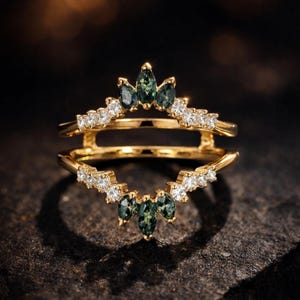 May include: A gold ring guard featuring two chevron-shaped bands connected at the sides. The bands are set with small, clear round stones and marquise-cut dark green stones arranged in a pointed, crown-like pattern at the centre of each band.