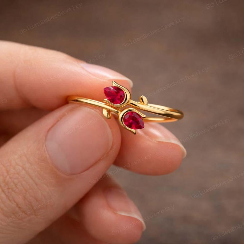 May include: A gold-colored ring featuring two pear-shaped, bright red gemstones. The ring's design incorporates a floral or botanical motif, with delicate curves and leaf-like accents. The ring is held in a hand.