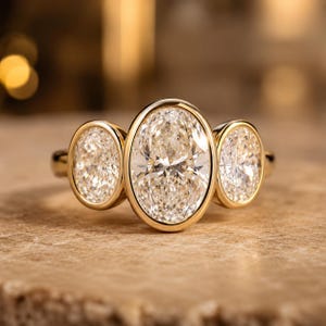 Puede incluir: Un anillo de oro con tres piedras claras de corte ovalado engastadas en bisel. La piedra central es más grande que las dos piedras ovaladas laterales. La banda de metal es de oro amarillo pulido y las piedras están dispuestas horizontalmente.