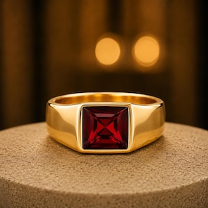May include: A gold signet ring featuring a square-cut, deep red gemstone set in the centre. The band is smooth and polished, with a wide, solid construction that tapers slightly towards the back. The gemstone displays a faceted surface.