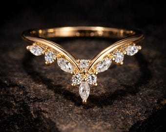 May include: A gold V-shaped ring featuring a central cluster of clear marquise and round-cut stones. The band follows a chevron silhouette, with additional marquise and round-cut stones set along the top edges, creating a symmetrical, pointed design.