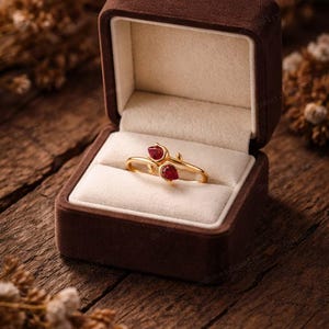 May include: A gold ring with two red gemstones, presented in a brown velvet jewelry box. The ring has a delicate, floral design. The box is open, revealing the ring on a cream-colored cushion, set against a rustic wooden background.