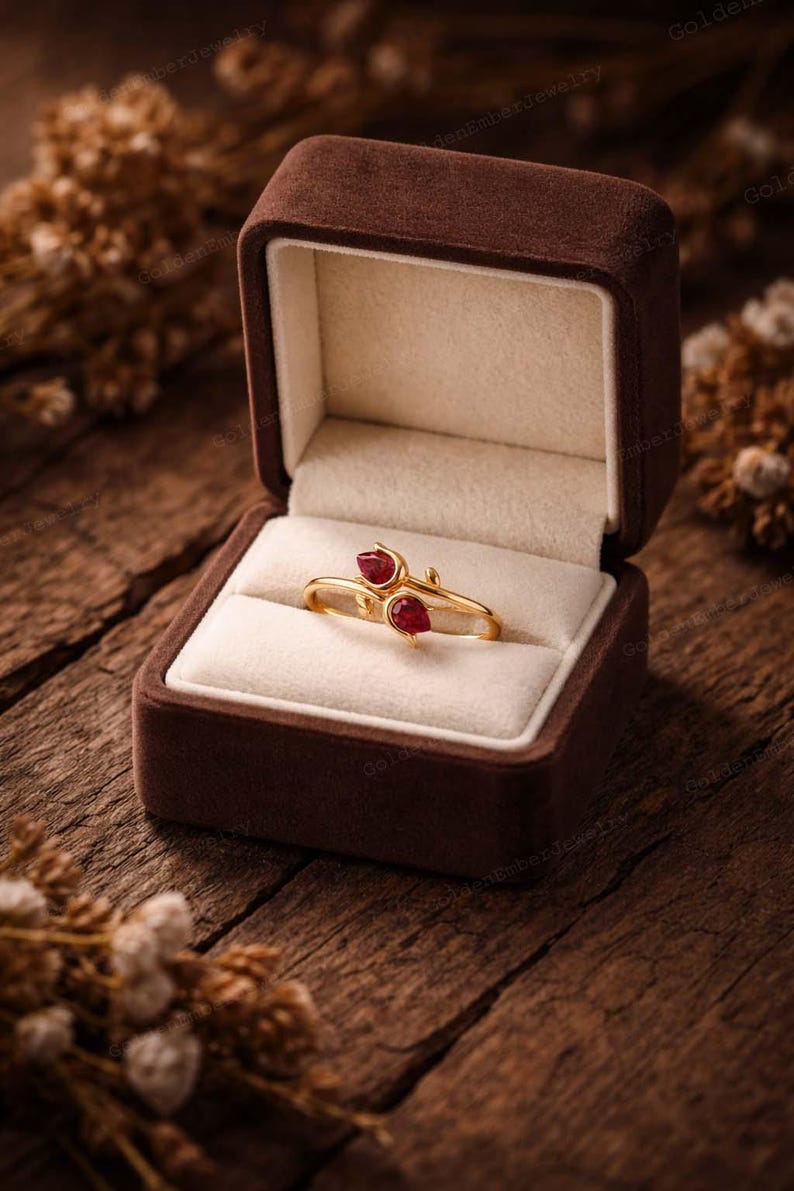 May include: A gold ring with two red gemstones, presented in a brown velvet jewelry box. The ring has a delicate, floral design. The box is open, revealing the ring on a cream-colored cushion, set against a rustic wooden background.
